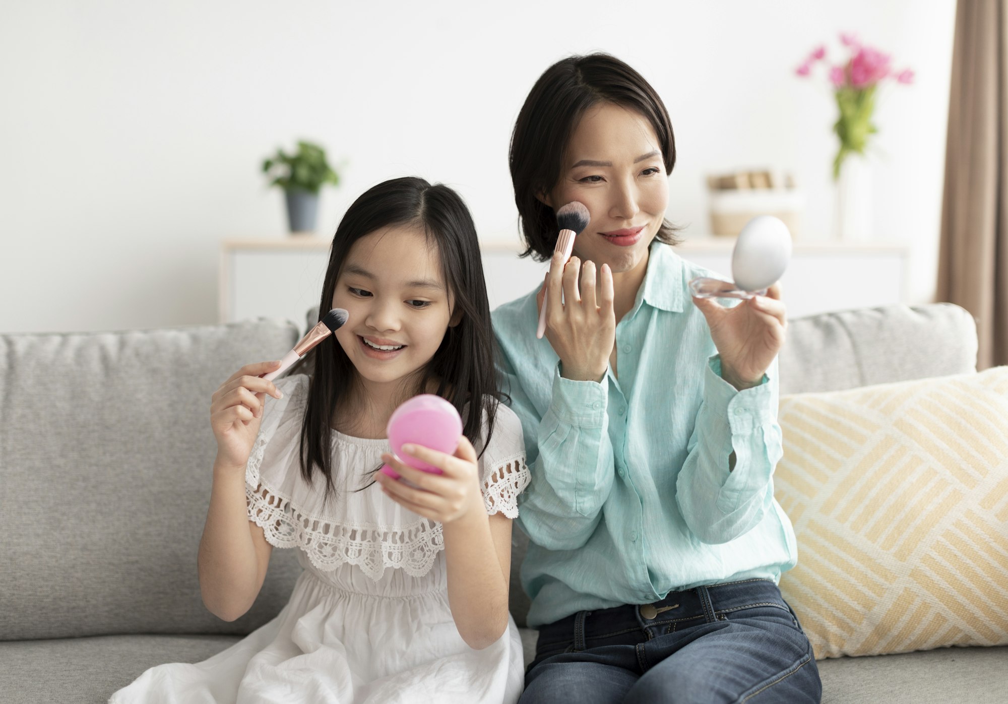 Happy mature Asian woman with her cute little granddaughter applying makeup, having fun at home