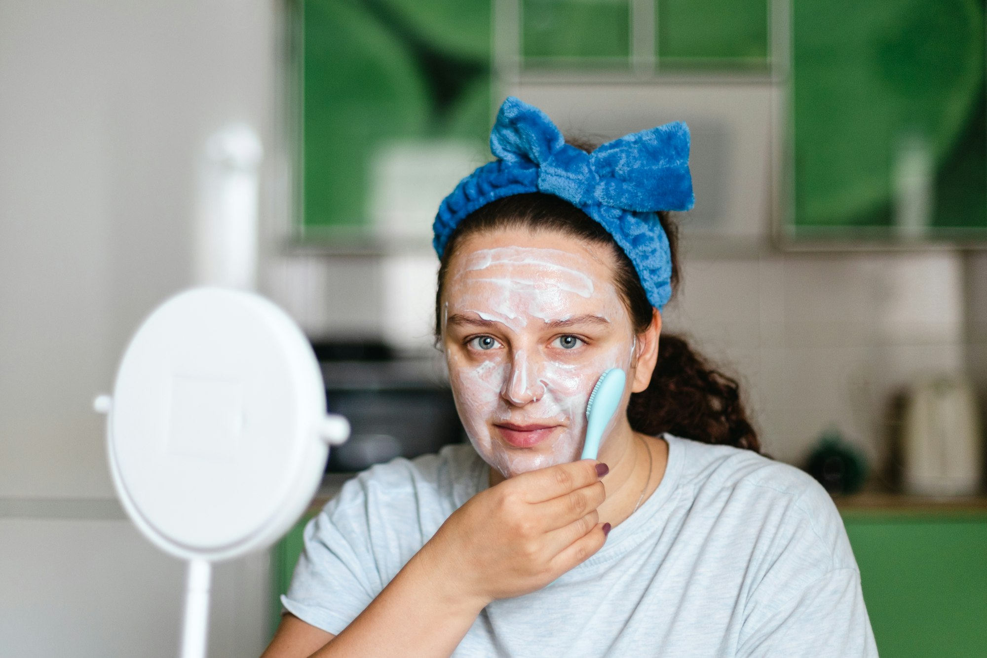 Woman massaging face skin with moisturizing mask with brush wearing blue bow on head looking at