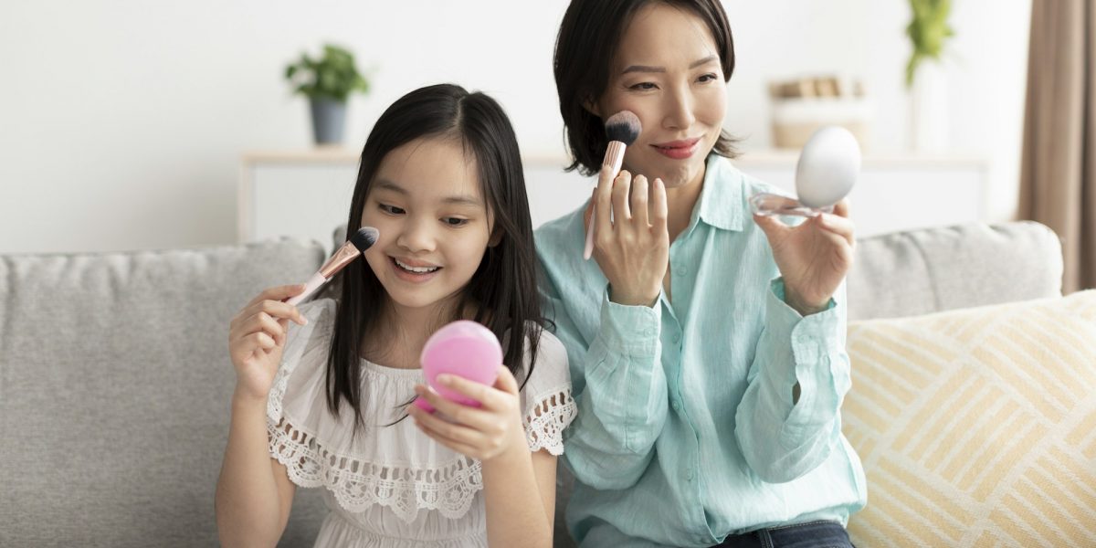 Happy mature Asian woman with her cute little granddaughter applying makeup, having fun at home