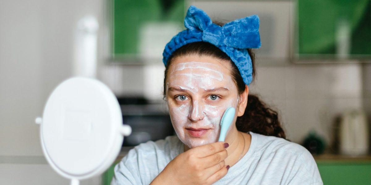 Woman massaging face skin with moisturizing mask with brush wearing blue bow on head looking at